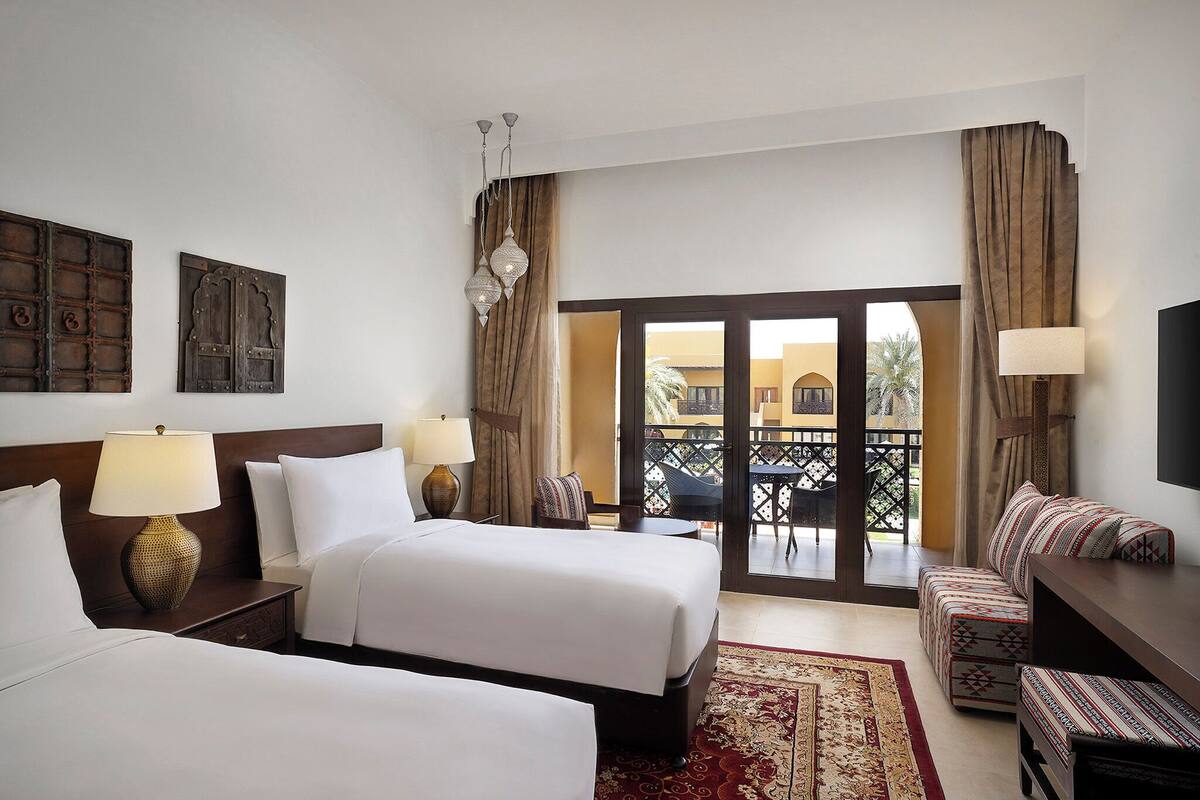 A hotel room with two beds, a sofa, a TV, a balcony with a view, and a patterned rug.