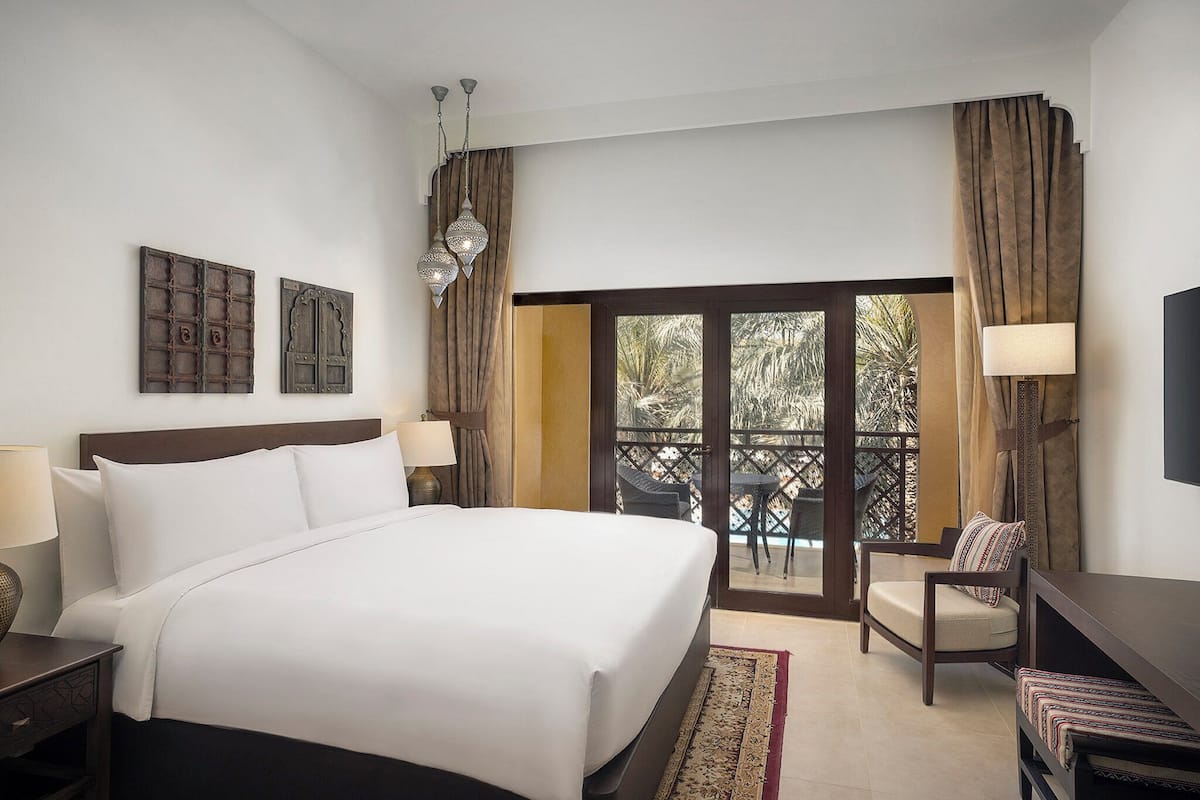 A hotel room with a large bed, a chair, a desk, and a balcony with a view.