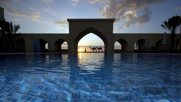 A swimming pool with a view of camels and a sunset through an archway. - Aldhafra Resort, Vignette Collection by IHG (Madinat Zayed)