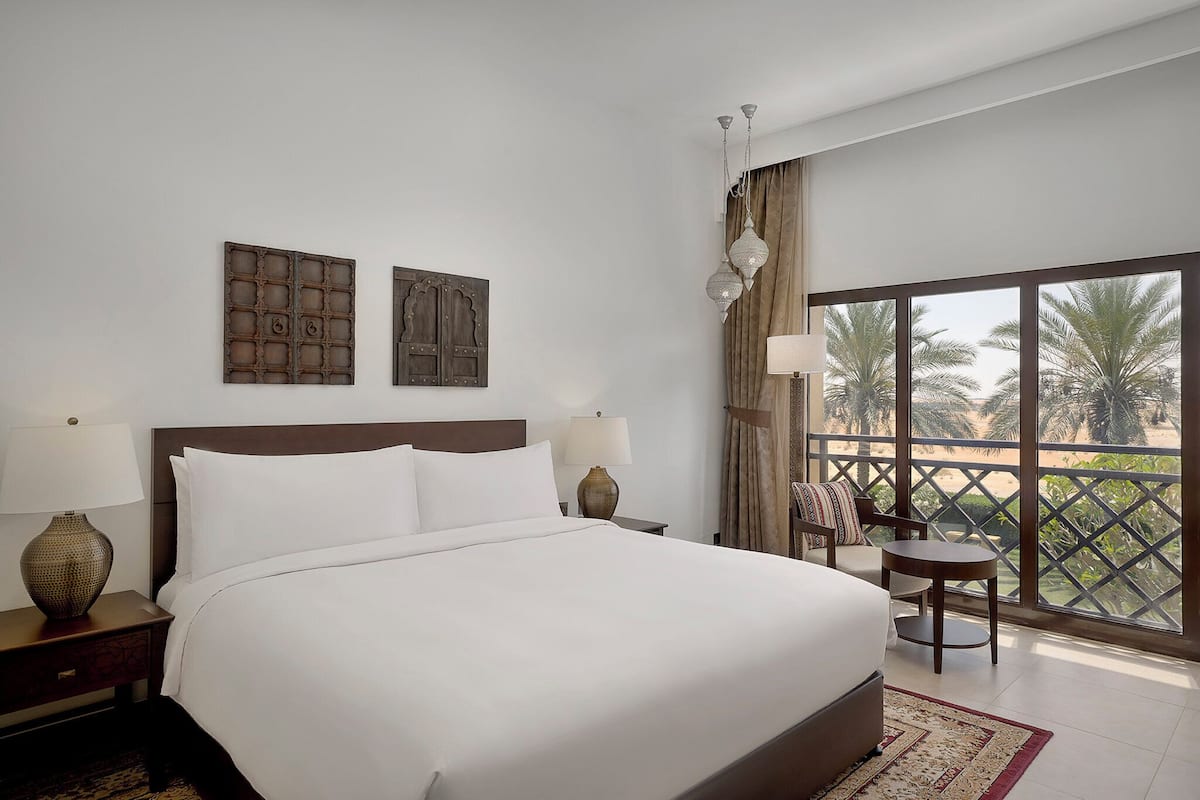 A hotel room with a large bed, two bedside tables with lamps, a view of palm trees through large windows, and a balcony with a chair and table.
