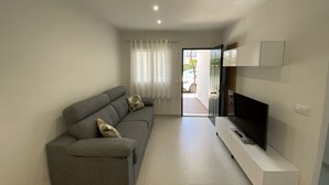 A modern living room with a grey sofa, a flat-screen TV on a white stand, and a door leading outside.