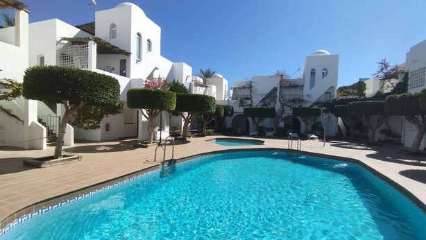 A swimming pool surrounded by white buildings with arched windows and a clear blue sky.