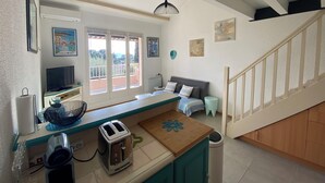Une cuisine équipée d’un grille-pain, d’une bouilloire et d’une petite table. Une fenêtre offre une vue sur l’extérieur. On aperçoit un escalier au fond.
