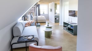 Un apartamento tipo loft moderno con cama alta, un sofá gris, una pequeña silla rosa, una mesa de centro y un televisor en una esquina.
