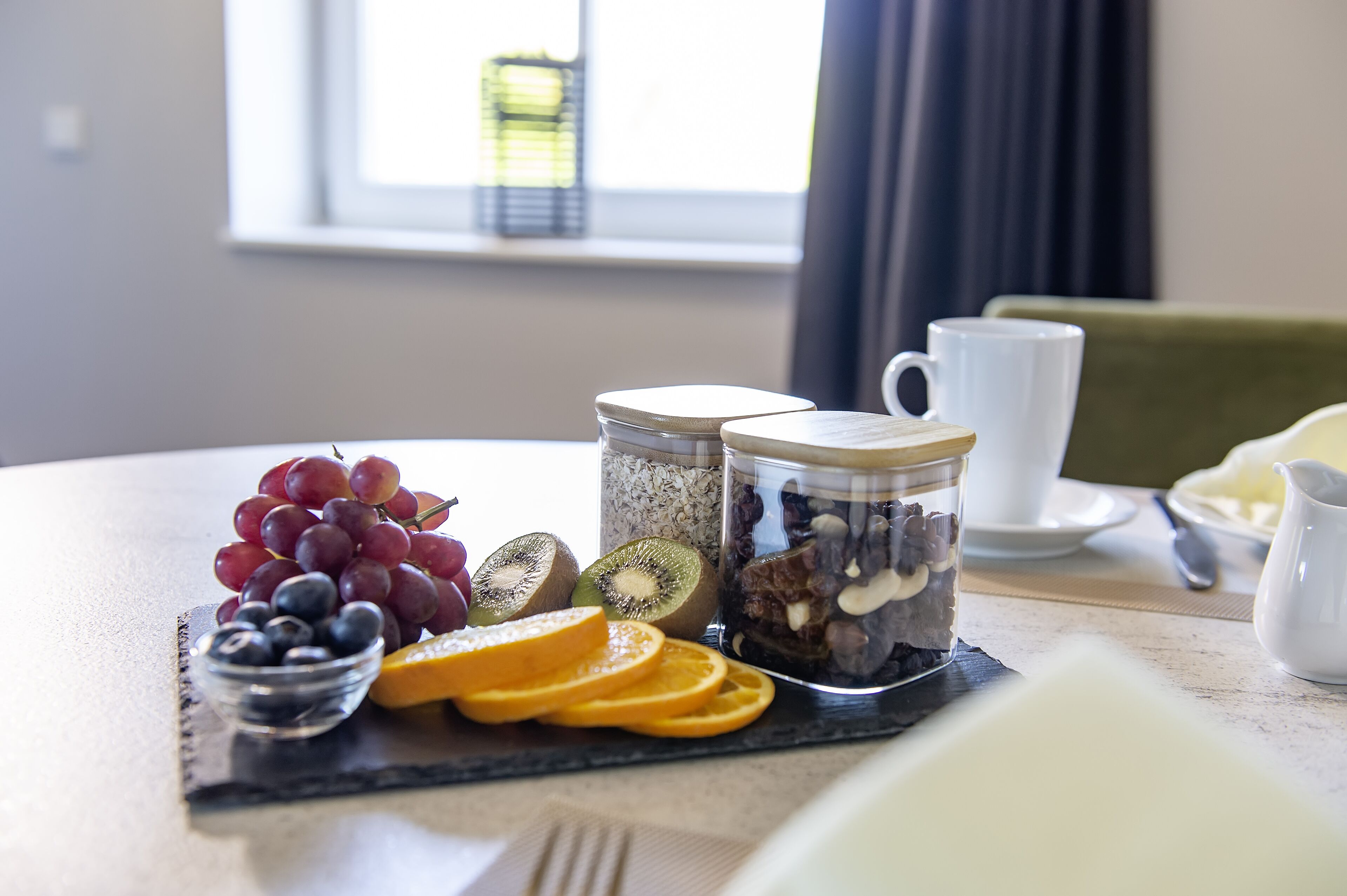 Una escena de desayuno con un bol de fruta, botes de nueces y semillas, y una taza sobre una mesa.