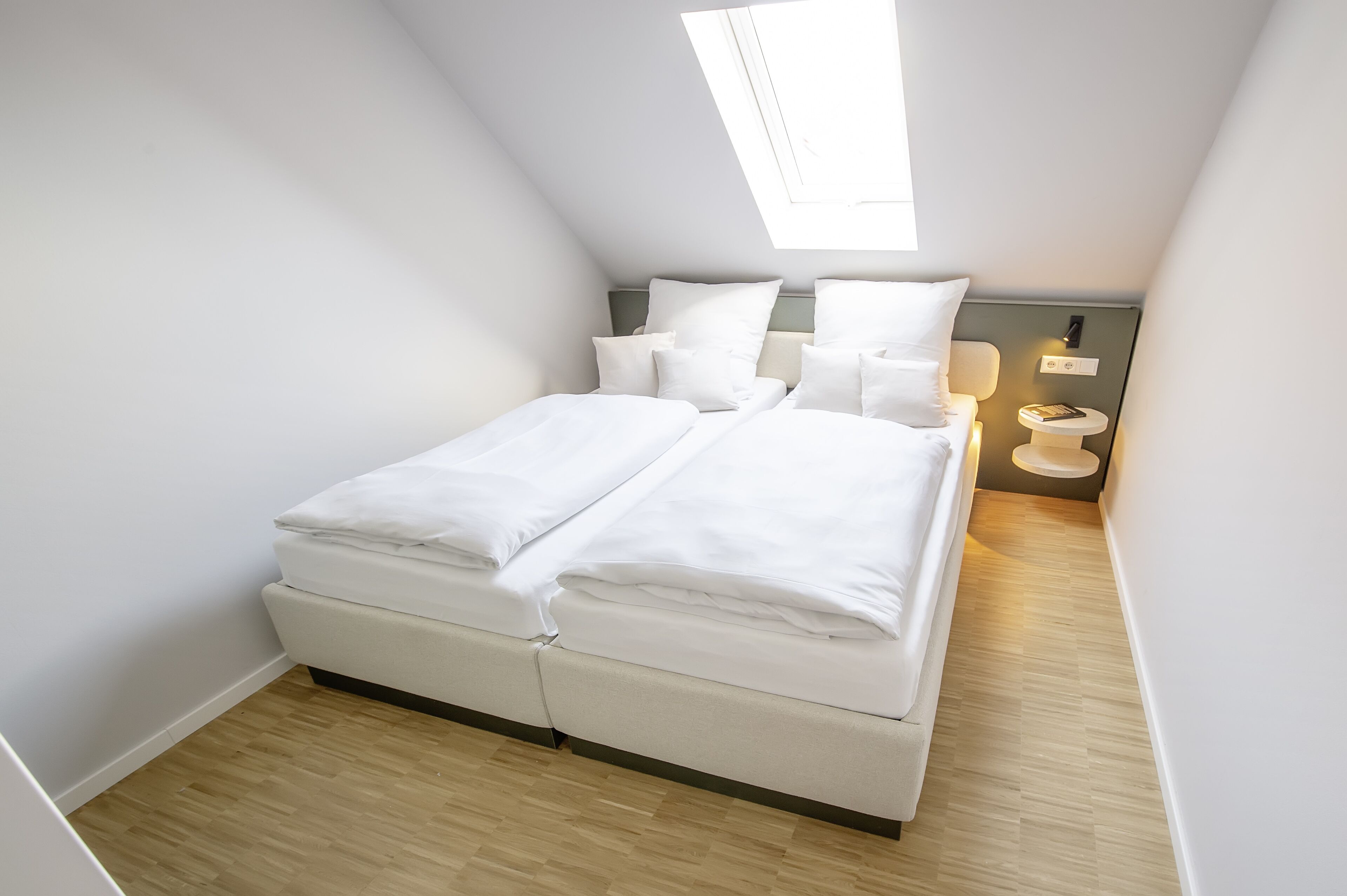 Une chambre à coucher moderne et bien éclairée, dotée d’une lucarne, d’un lit avec du linge de lit blanc, d’une table de chevet et d’une petite lampe de chevet.