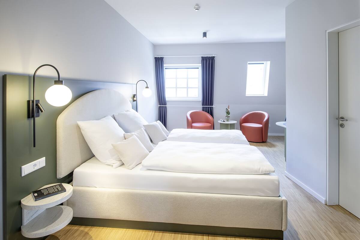 Habitación de hotel moderna con una cama grande, una mesita de noche, una lámpara de lectura y un ventanal con cortinas.