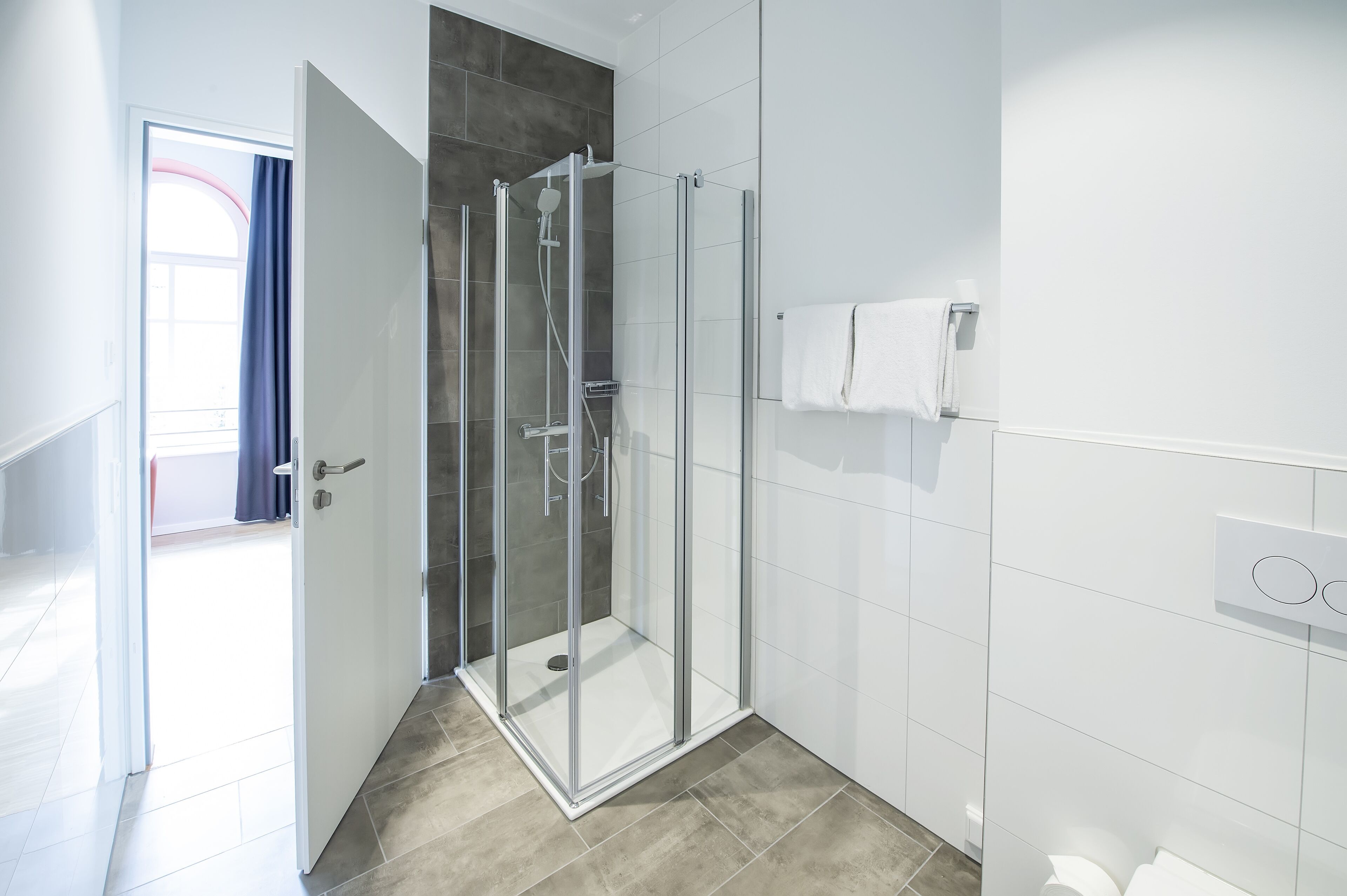 Un baño moderno con cabina de ducha de vidrio, paredes con azulejos blancos y un perchero visible.