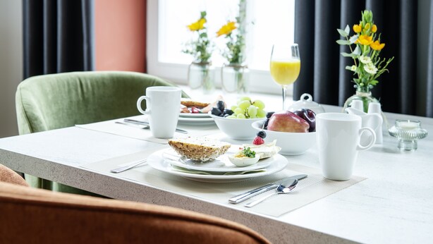 Un desayuno preparado para dos personas, con una mesa que incluye tostadas, huevos, fruta, un vaso de zumo de naranja y un florero con flores.
