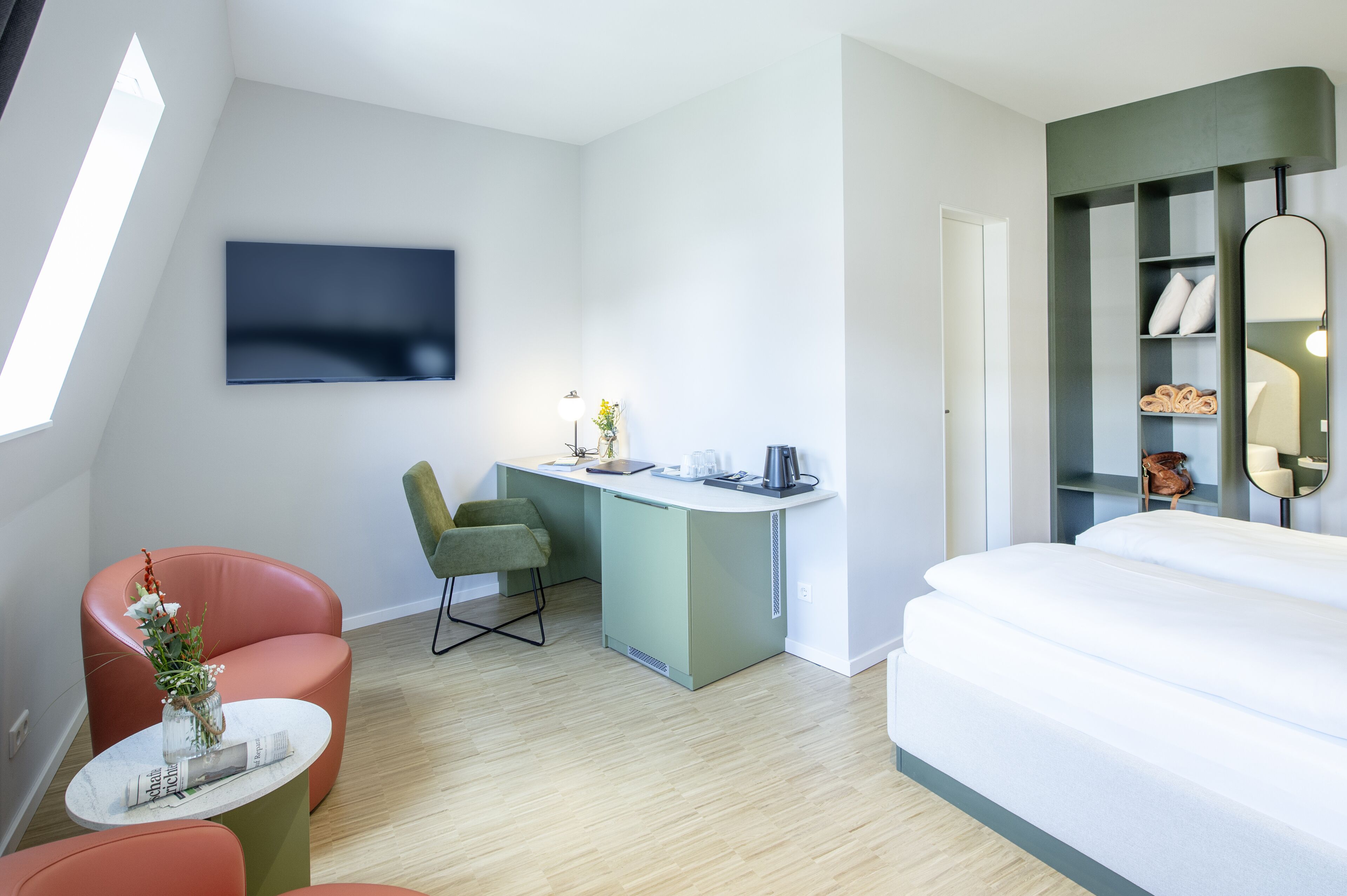 Ein modernes Hotelzimmer mit einem Bett, einem Schreibtisch mit Stuhl, einem an der Wand befestigten Fernseher und einem kleinen Tisch mit einer Vase.
