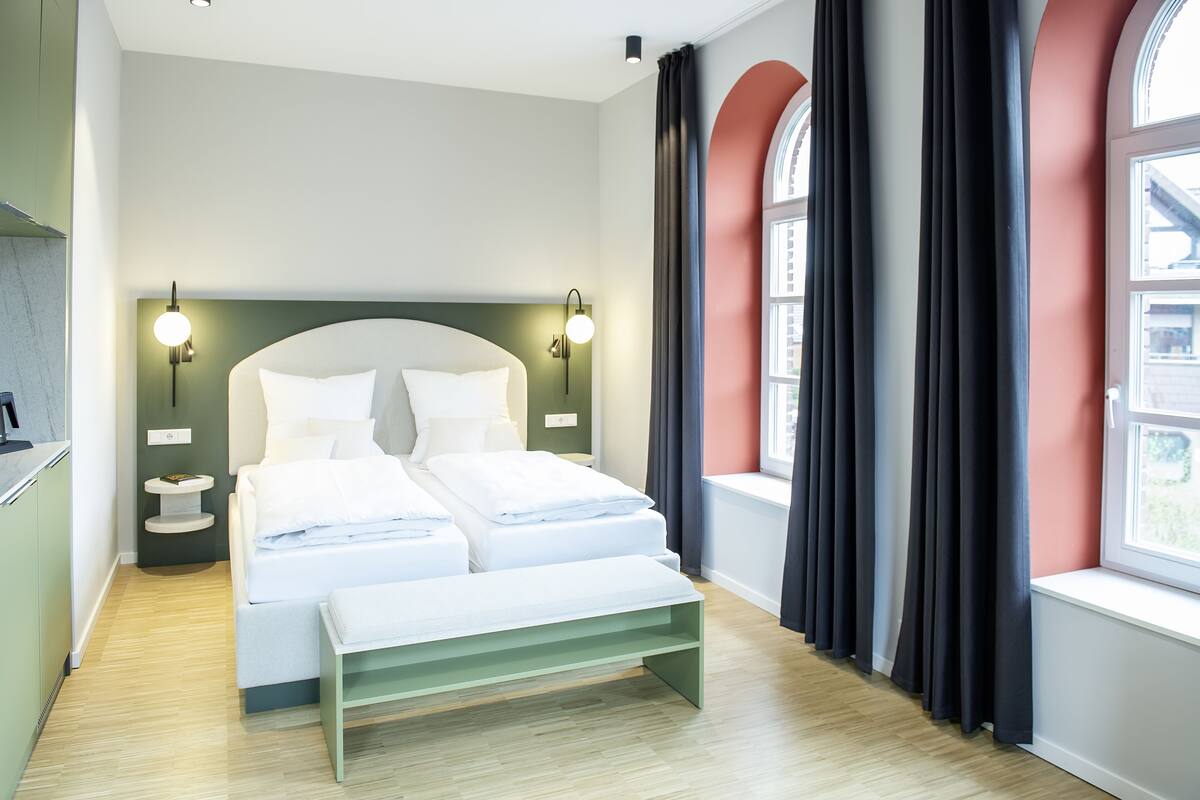 Una habitación de hotel moderna con una cama grande, dos mesitas de noche, un banco y una ventana con cortinas.