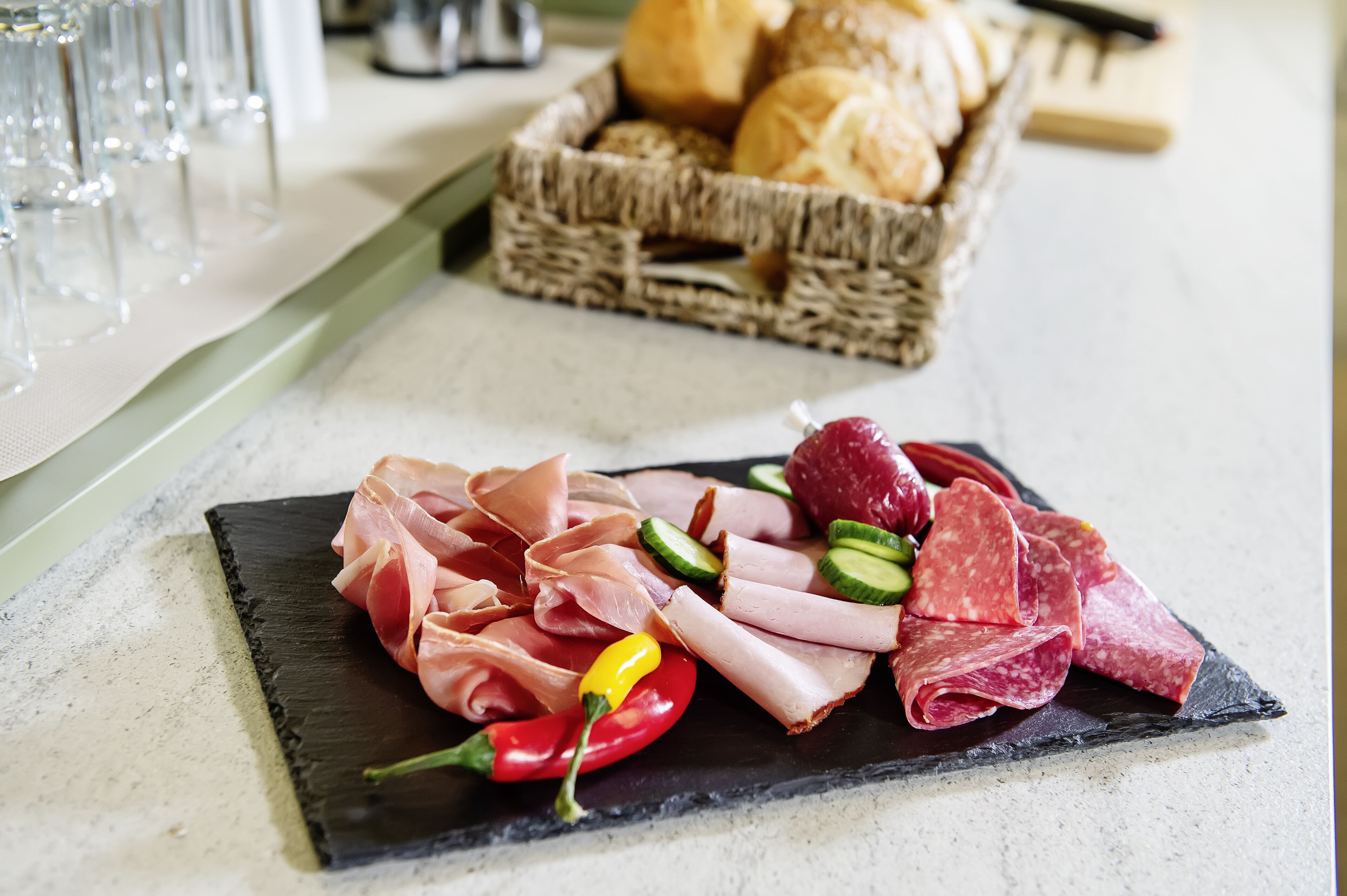 Una tabla de embutidos con diversos tipos de carne y verduras sobre una base de pizarra, una cesta de pan al fondo, y una mesa dispuesta con vasos.