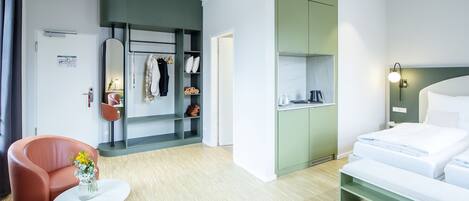Une chambre d’hôtel moderne comprenant un lit, un coin salon avec un fauteuil et une petite table, une armoire et une kitchenette.