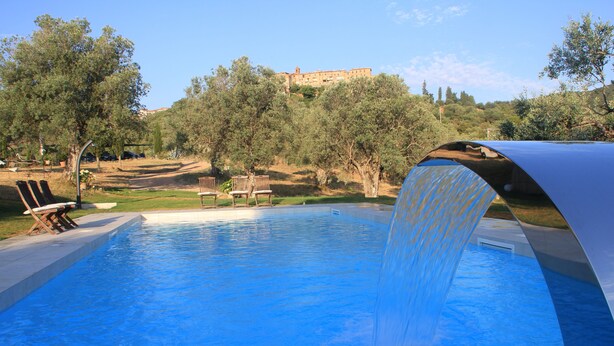 Une piscine agrémentée d’une cascade, entourée d’arbres et avec un bâtiment visible au loin.