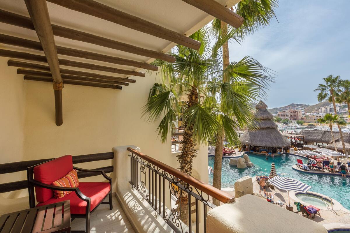 Un balcón con barandilla de madera, una silla con cojín rojo y vistas a una zona de piscina con sombrillas y tumbonas.