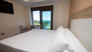 A hotel room with a large bed, a TV mounted on the wall, a balcony with a view of the ocean, and a small table with chairs. - A Ballenera de Caneliñas (Cee)