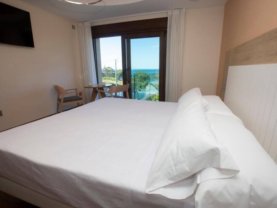 A hotel room with a large bed, a TV mounted on the wall, a balcony with a view of the ocean, and a small table with chairs.