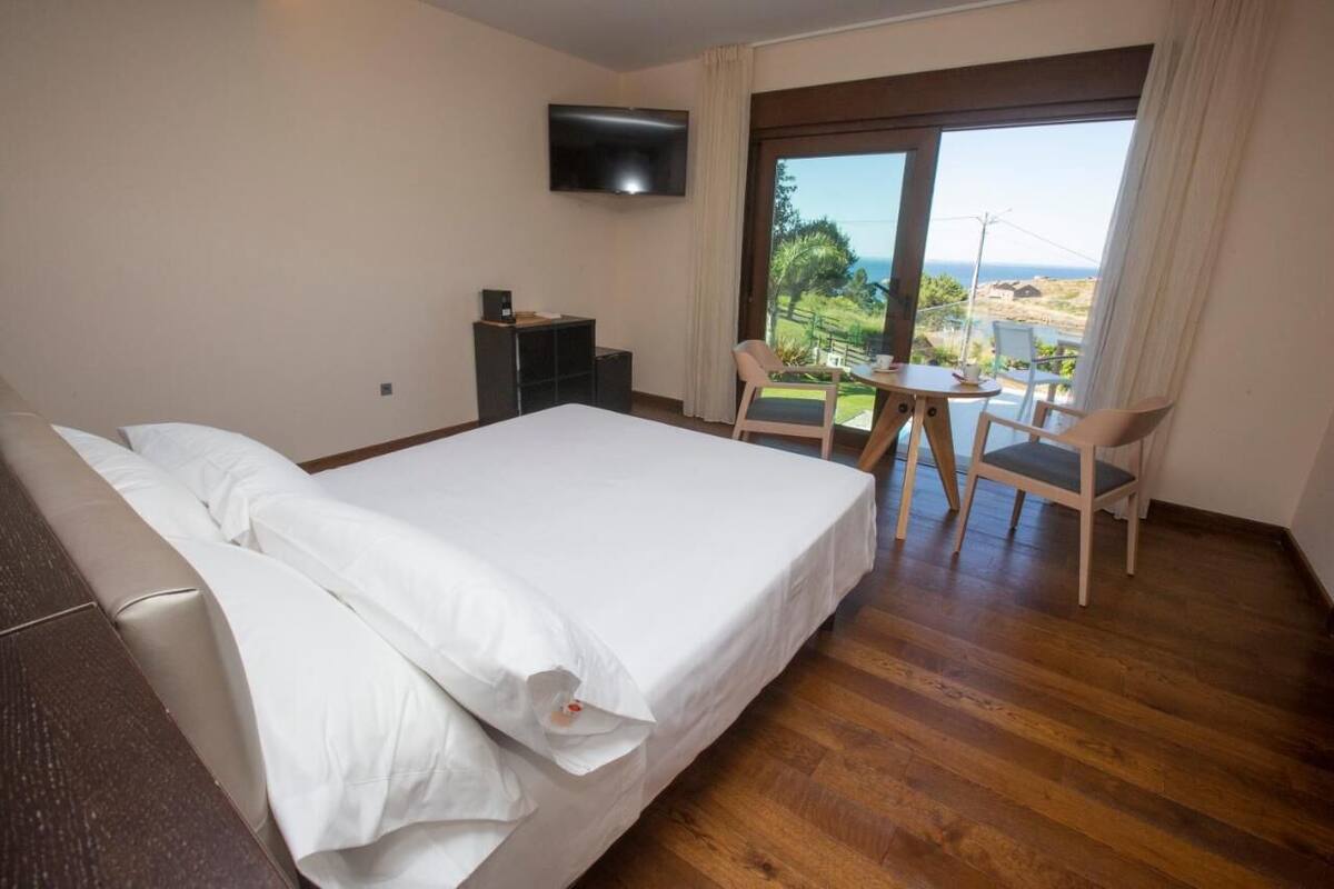 Une chambre d’hôtel avec un grand lit, un téléviseur fixé au mur, un balcon avec une table et des chaises, et une vue sur la mer.
