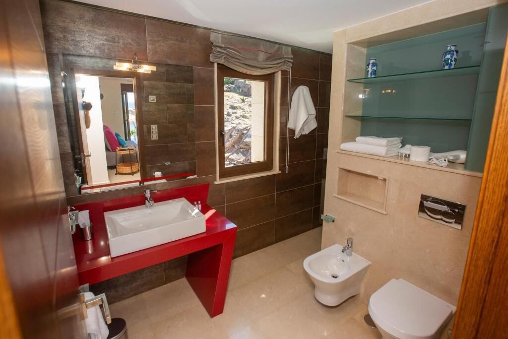 A bathroom with a red vanity, a white sink, a bidet, and a glass shelf with toiletries.
