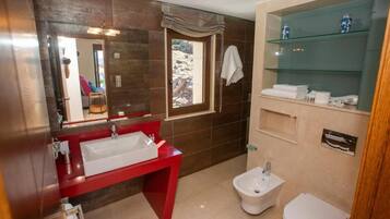 A bathroom with a red vanity, a white sink, a bidet, and a glass shelf with toiletries.