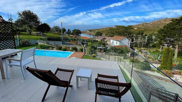 A balcony with two wooden chairs and a small table, overlooking a swimming pool and a scenic view of a coastal town. - A Ballenera de Caneliñas (Cee)
