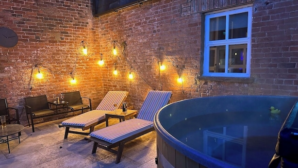 Une terrasse extérieure accueillante, avec des fauteuils confortables, un jacuzzi et des guirlandes lumineuses disposées le long d’un mur de briques.