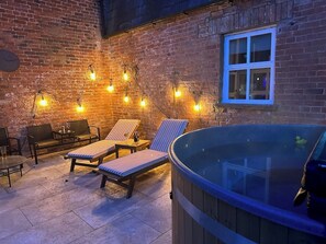 A cozy outdoor patio with lounge chairs, a hot tub, and string lights against a brick wall.