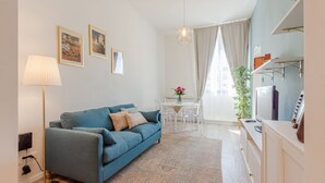 A living room with a blue sofa, a floor lamp, a dining table with chairs, and a kitchen area with a TV and cabinets. - Belvilla by OYO Maison (Fucecchio)