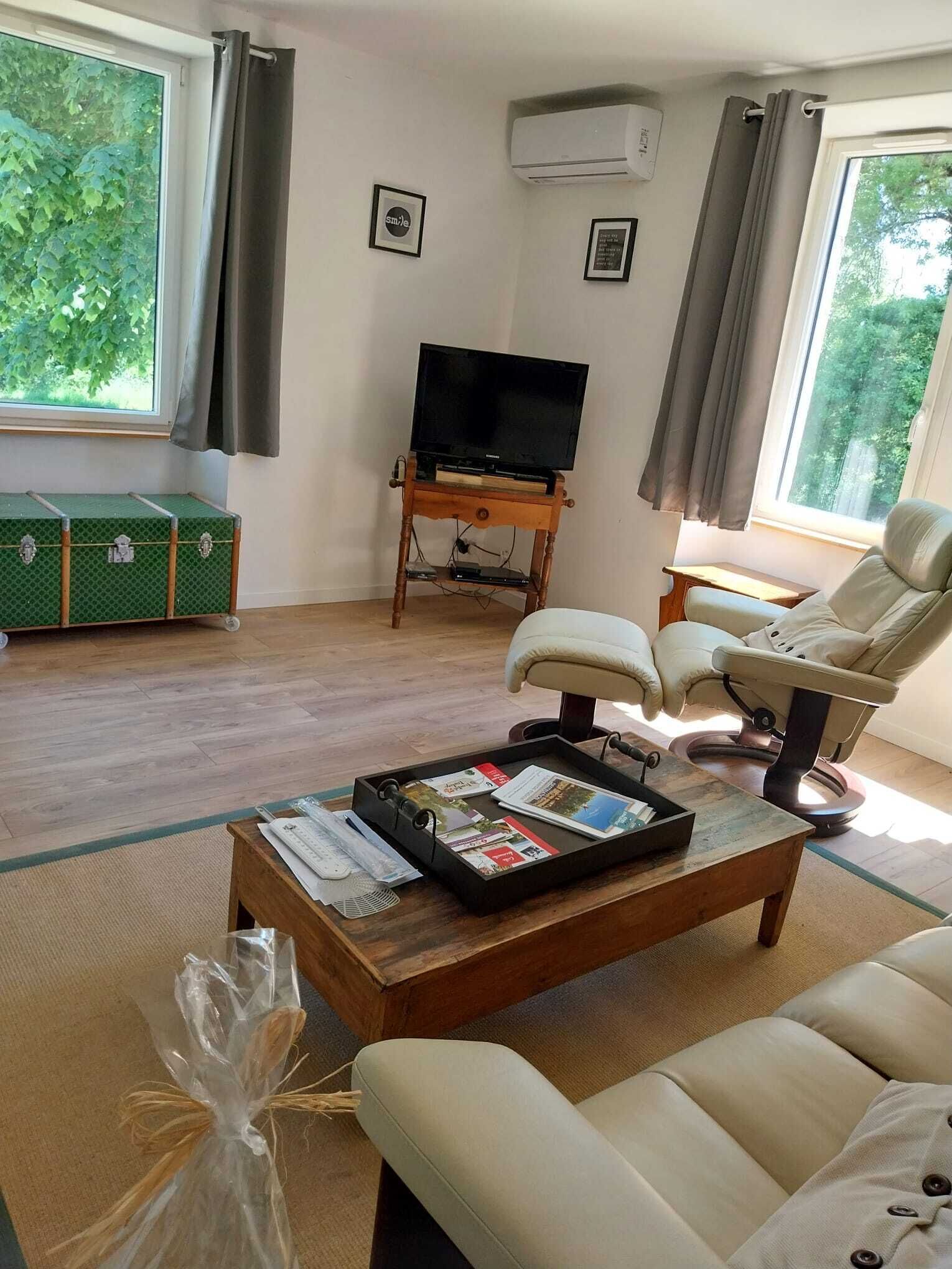 A living room with a sofa, a recliner, a coffee table with magazines, a television on a wooden stand, and a green suitcase.