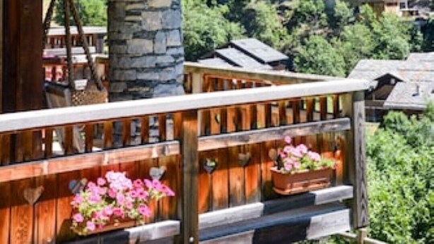 A wooden balcony with flower planters, a stone pillar, and a view of a mountainous village.