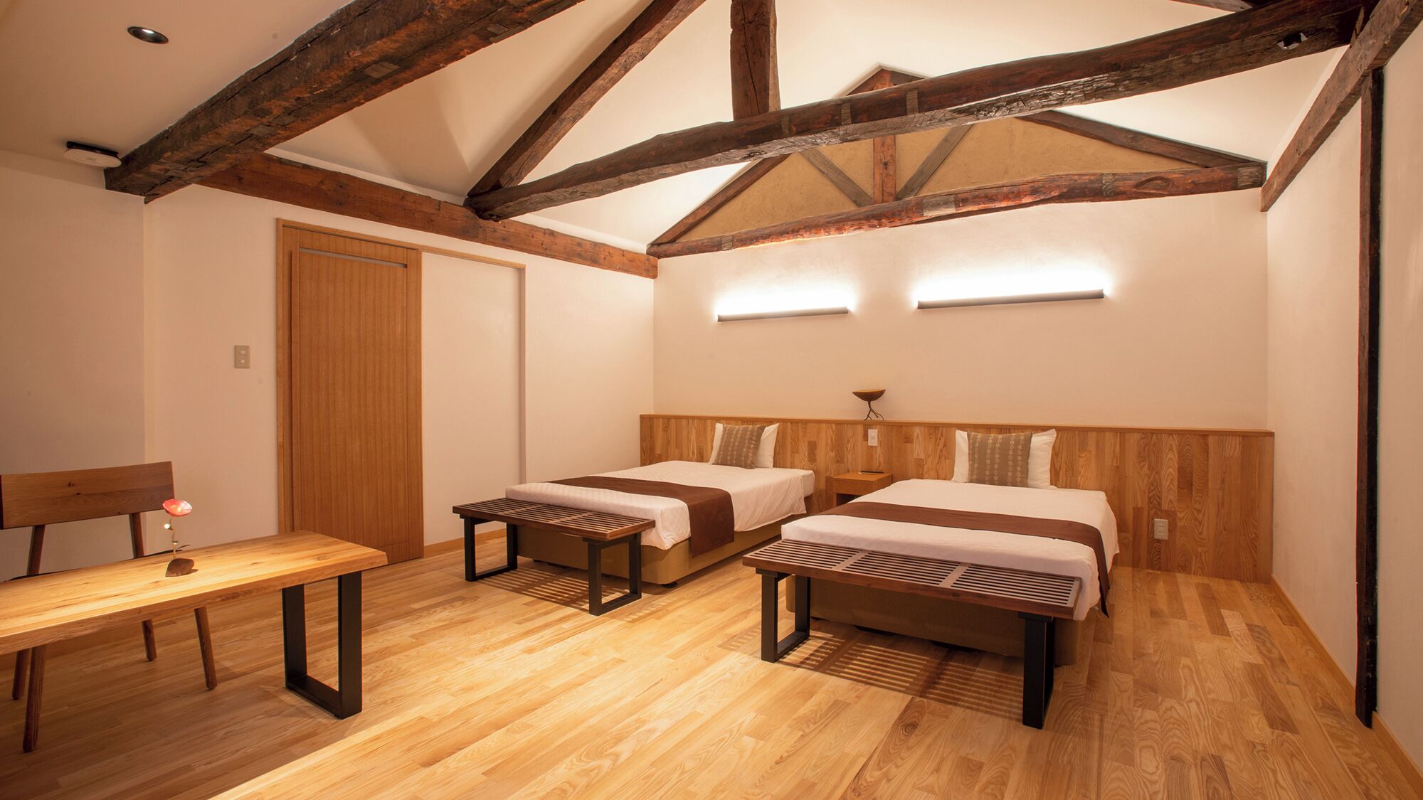 A room with two beds, a wooden bench, a desk, and a ceiling with exposed beams.