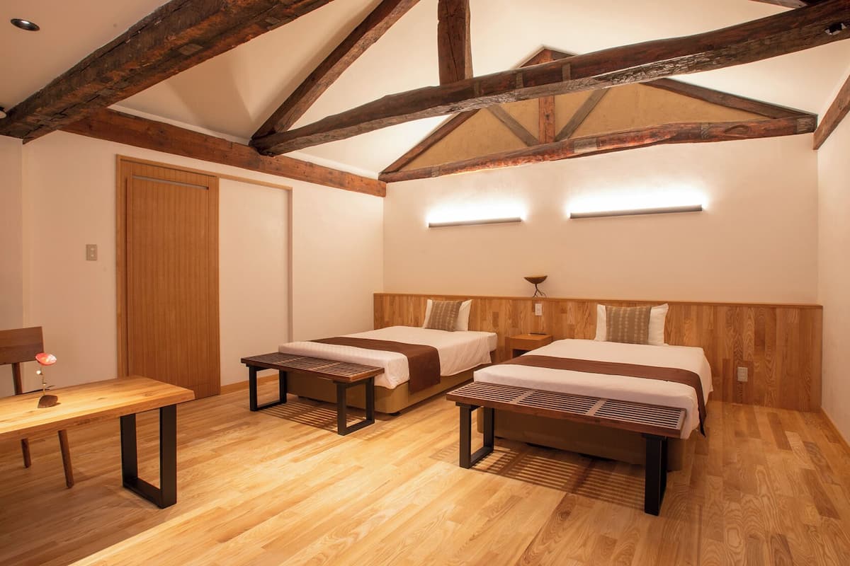 Ein Zimmer mit zwei Betten, einer Holzbank, einem Schreibtisch und einer Decke mit sichtbaren Holzbalken.