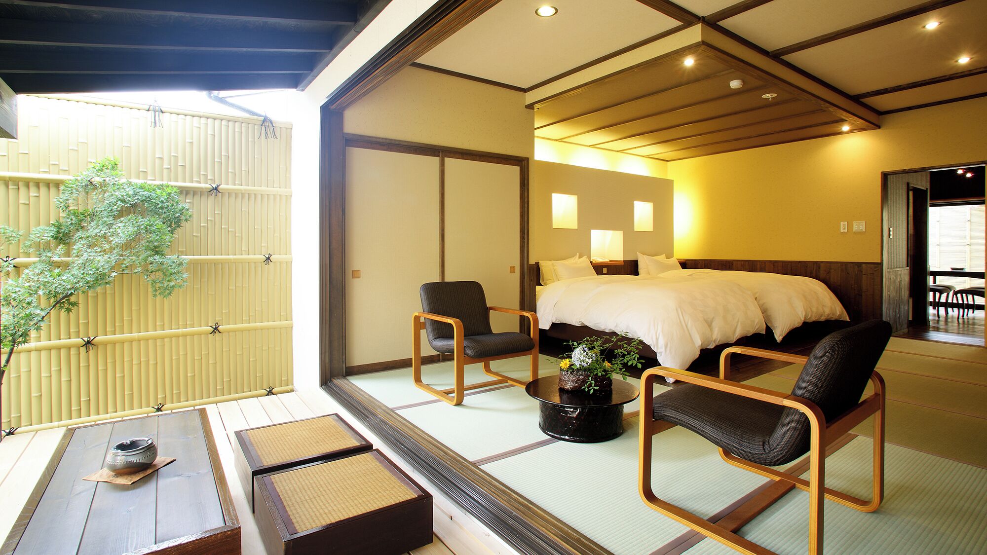 A traditional Japanese-style room with tatami flooring, a bed, two chairs, a small table with a bowl, and a potted plant.