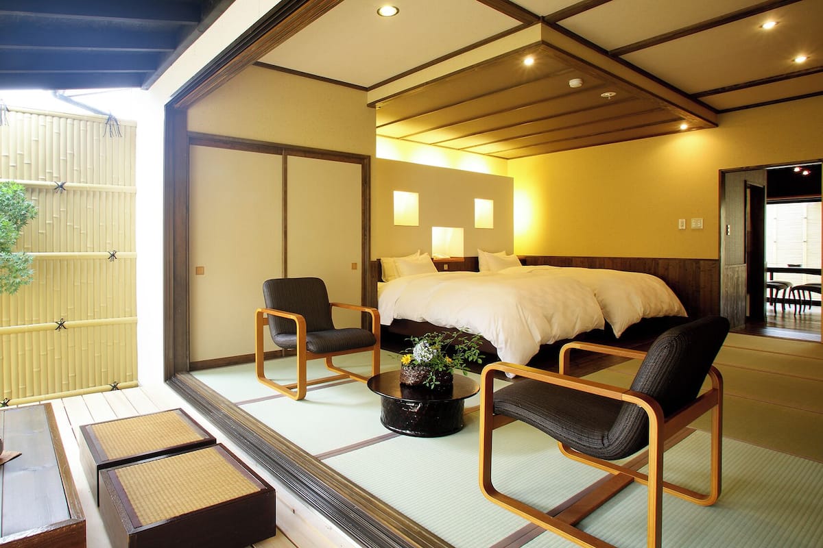 Ein traditionell japanisch gestalteter Raum mit Tatami-Böden, einem Bett, zwei Stühlen, einem kleinen Tisch mit einer Schale und einer Topfpflanze.