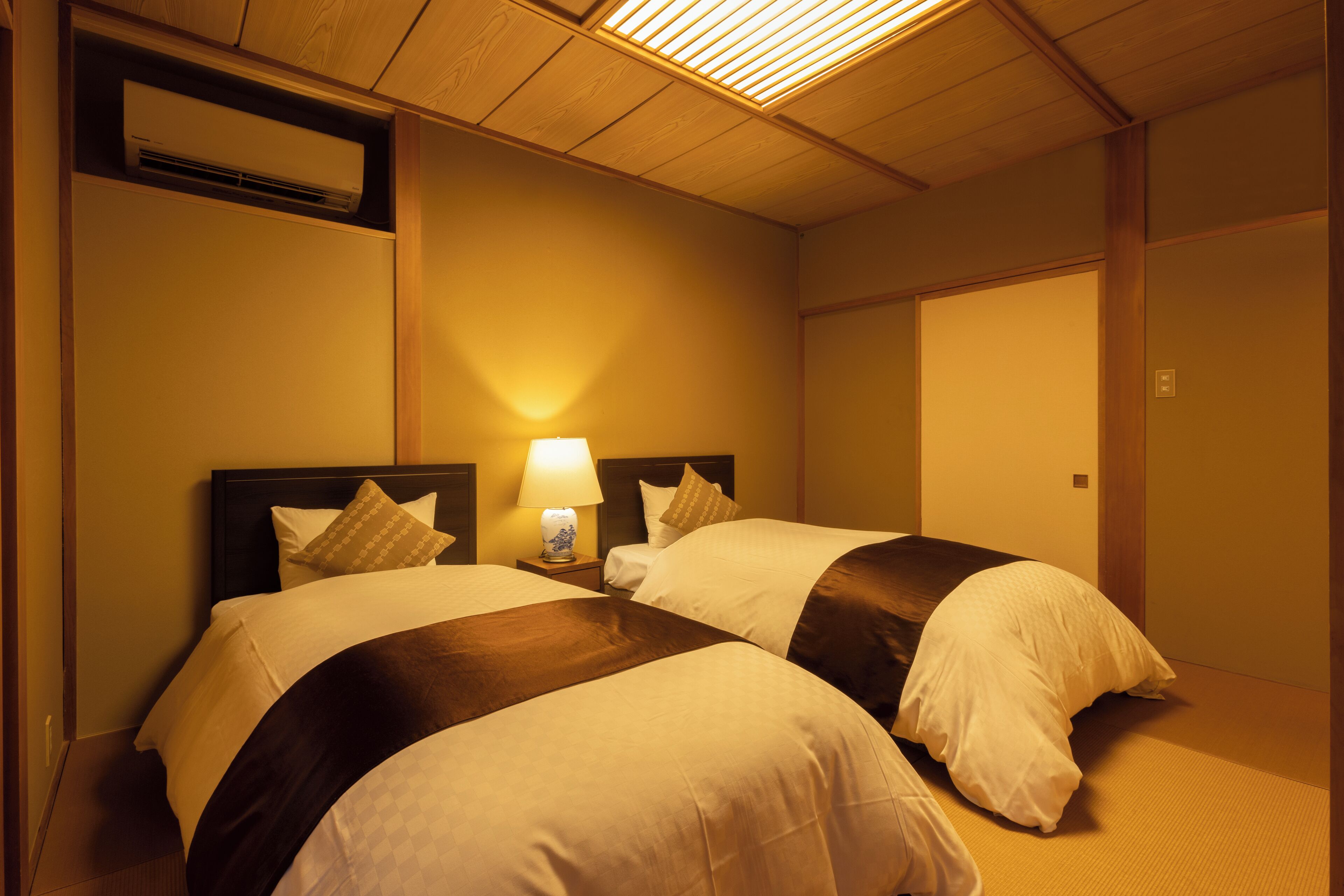 A hotel room with two beds, a nightstand with a lamp, and a wall-mounted air conditioner.