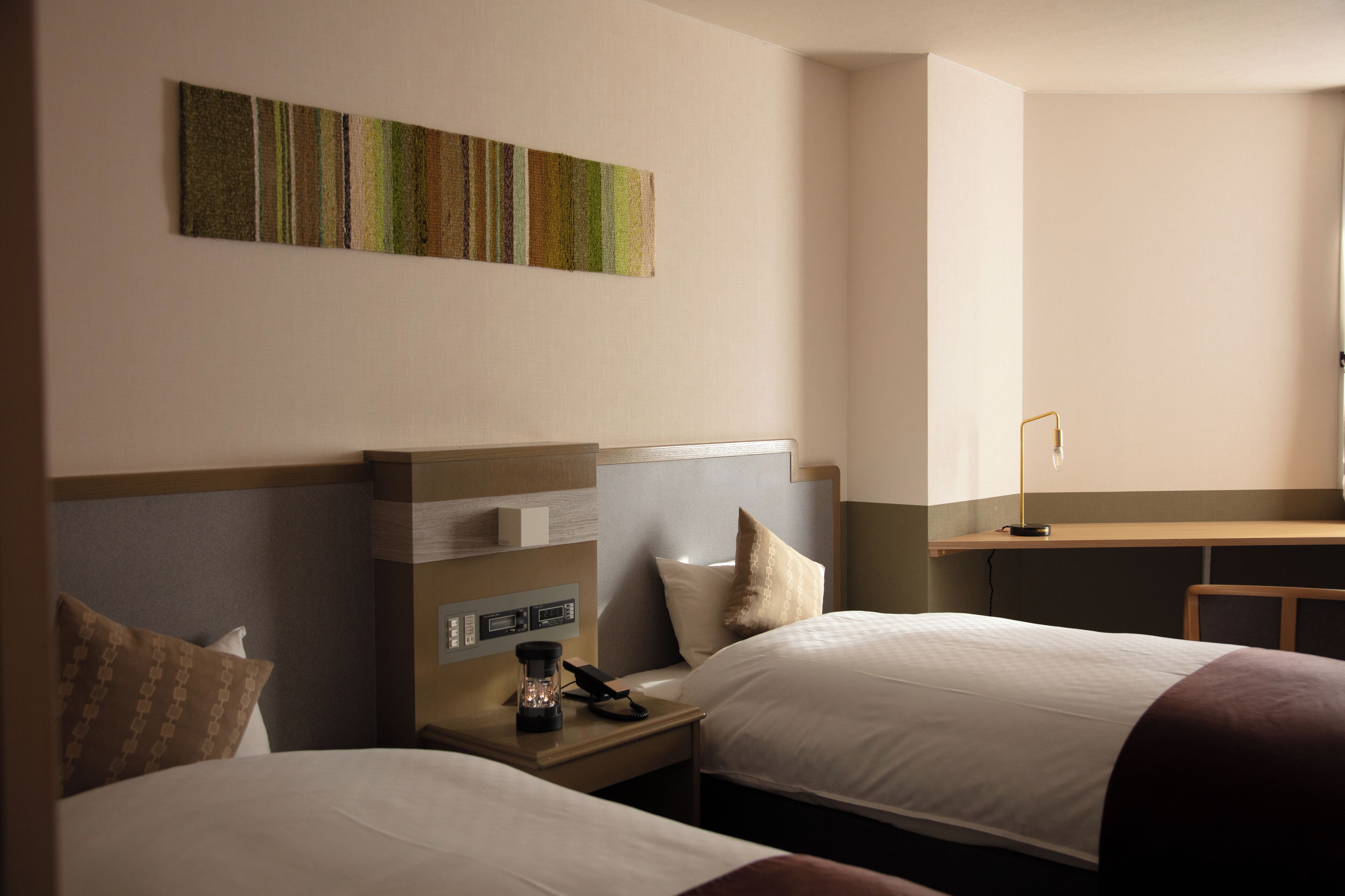 A hotel room with two beds, a desk, a lamp, and a wall-mounted artwork.