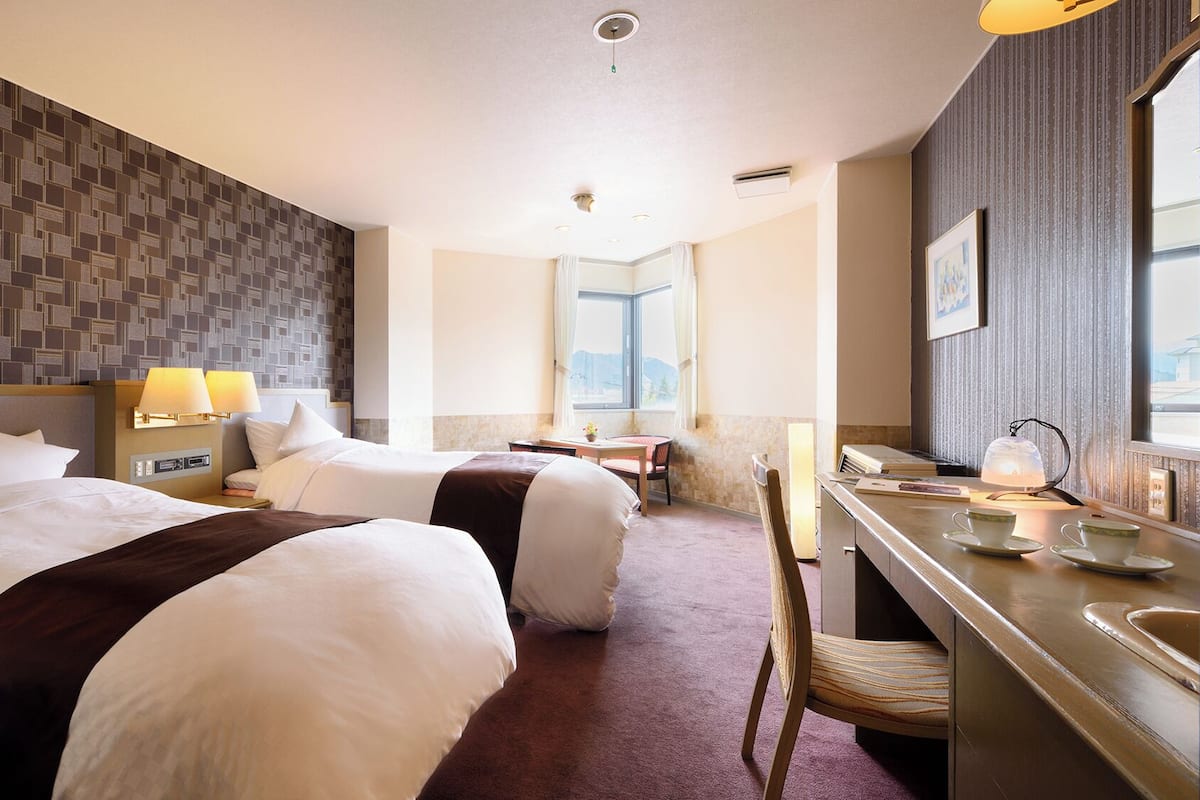 Ein Hotelzimmer mit zwei Betten, einem Schreibtisch, einem Stuhl, einem Fenster und Blick ins Freie.