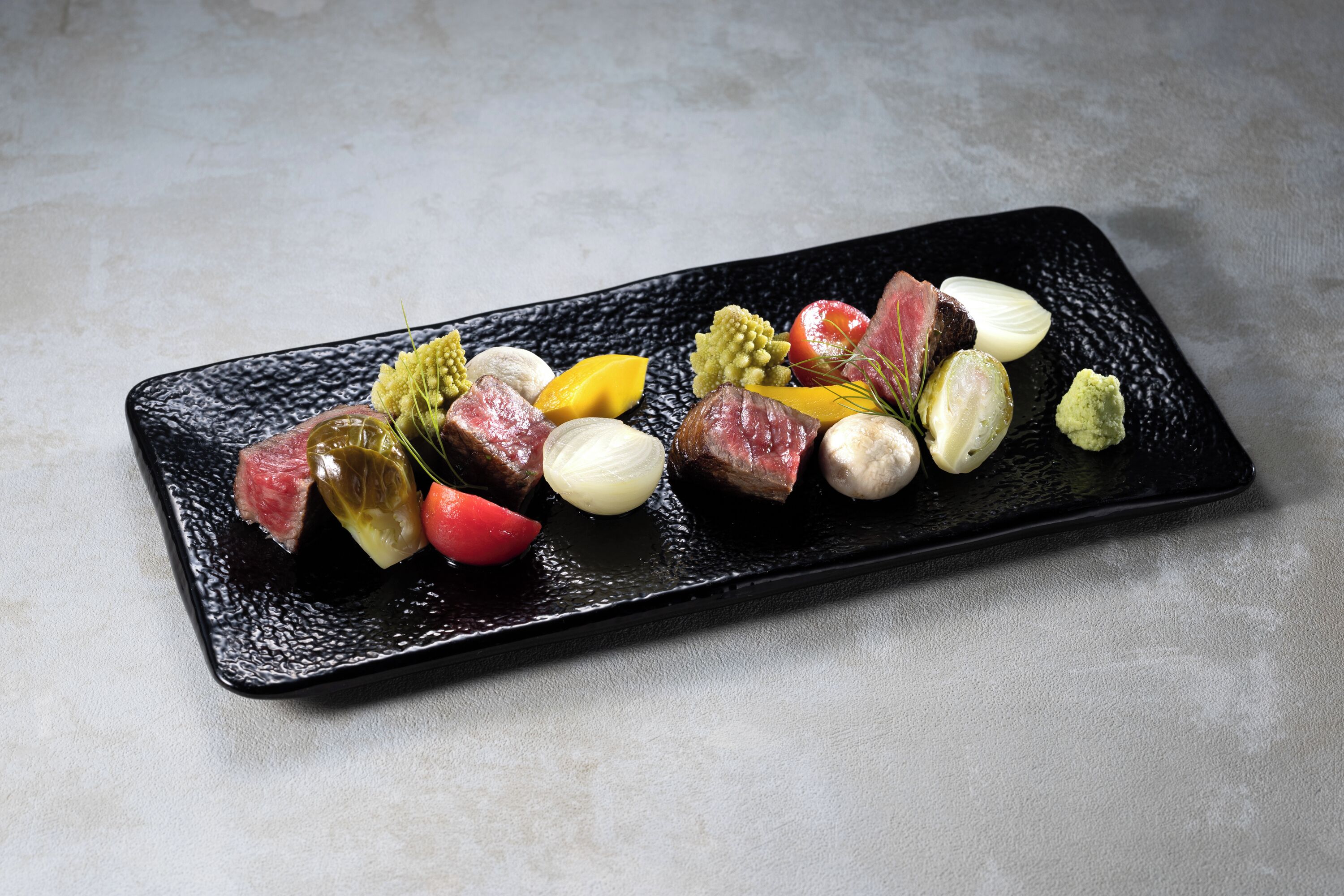 Un plato rectangular negro con diversos alimentos, incluyendo carne, verduras y posiblemente algunos condimentos.