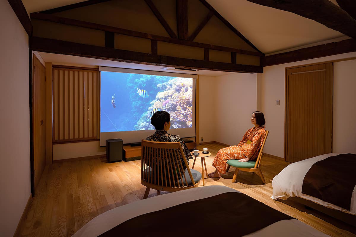 Ein Zimmer mit einem großen Bildschirm, der eine Unterwasserwelt zeigt, Holzbalken an der Decke, einem Bett und einer Sitzecke mit kleinem Tisch und Stühlen.