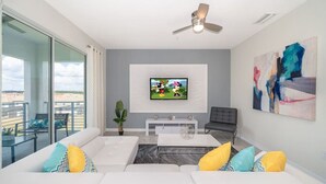 A modern living room with a flat-screen TV, a ceiling fan, and a balcony with outdoor seating. - 3121-406 · Modern Apartment with Privilage Location & View (Kissimmee)