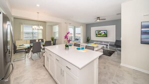 A modern kitchen with a central island, dining area with chairs and a table, a television mounted on the wall, and a ceiling fan. - 3121-406 · Modern Apartment with Privilage Location & View (Kissimmee)