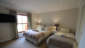 Une chambre d’hôtel avec deux lits, un téléviseur fixé au mur, une fenêtre avec des rideaux et une table de chevet avec une lampe et un vase de fleurs.