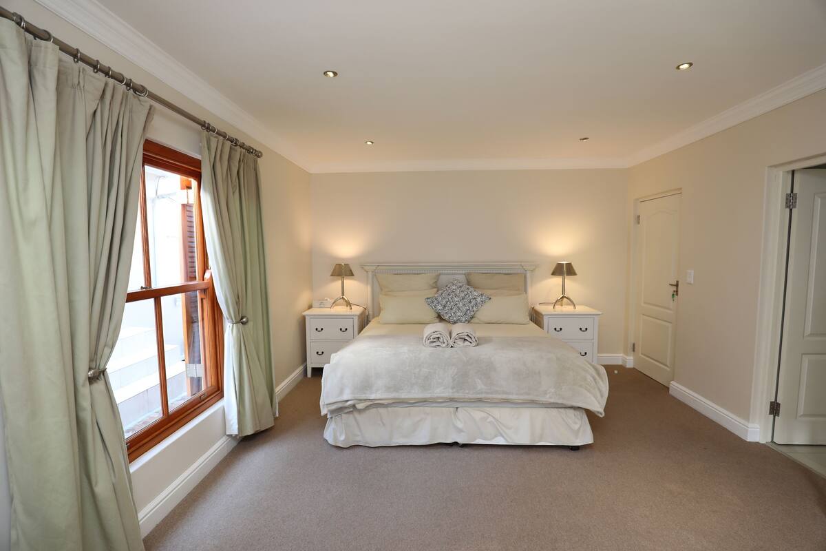 A bedroom with a large bed, two bedside tables with lamps, a window with curtains, and a door.