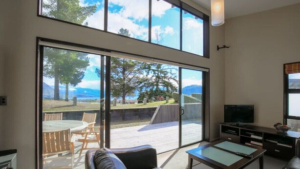 A spacious living room with a large sliding glass door leading to an outdoor patio, a leather sofa, a coffee table, and a flat-screen TV. - Anderson Heights | Spacious Stay with lake views (Wānaka)