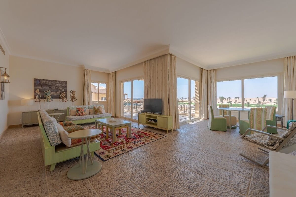 Uma sala de estar ampla com uma televisão grande, sofás verdes e vista para a praia.