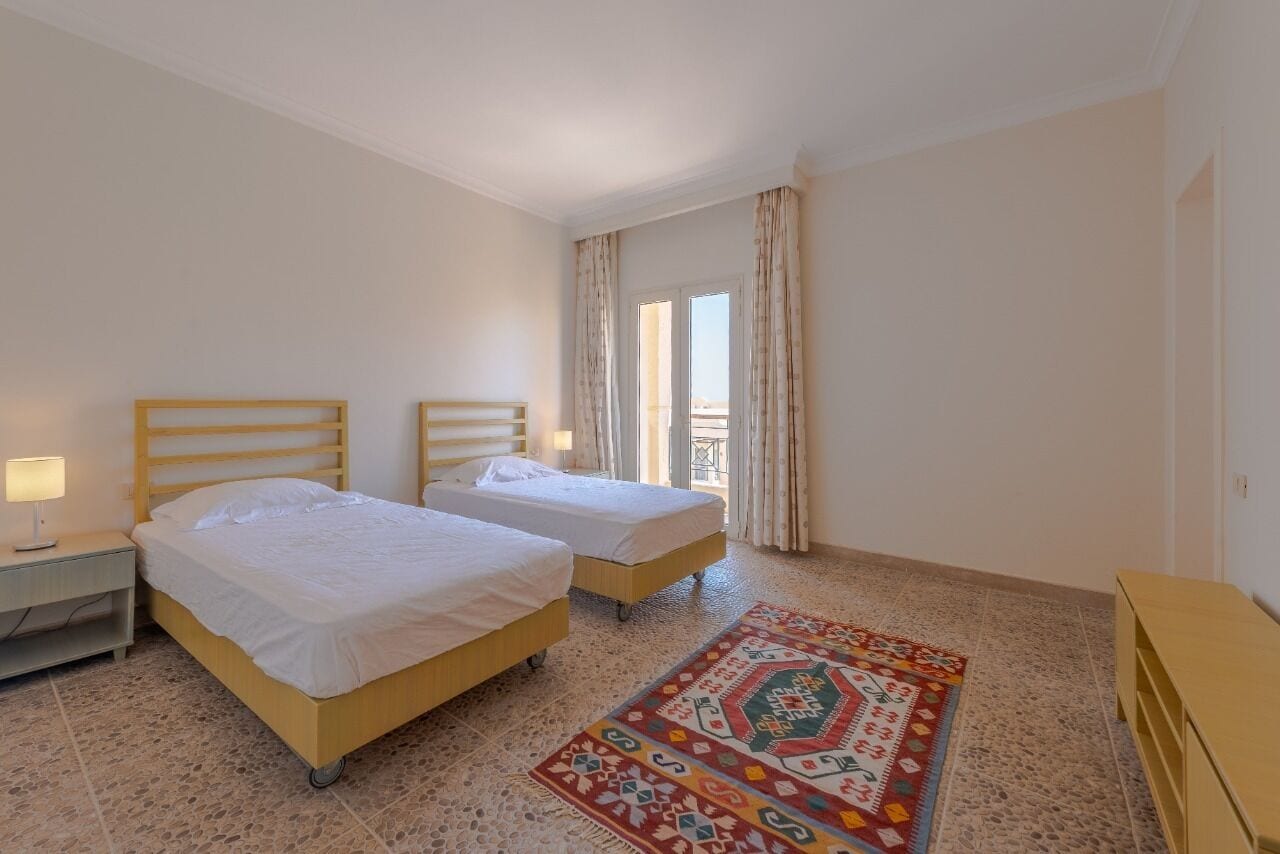 Une chambre d’hôtel avec deux lits simples, une petite table de chevet avec une lampe, un tapis coloré et une vue sur l’extérieur par une fenêtre avec des rideaux.