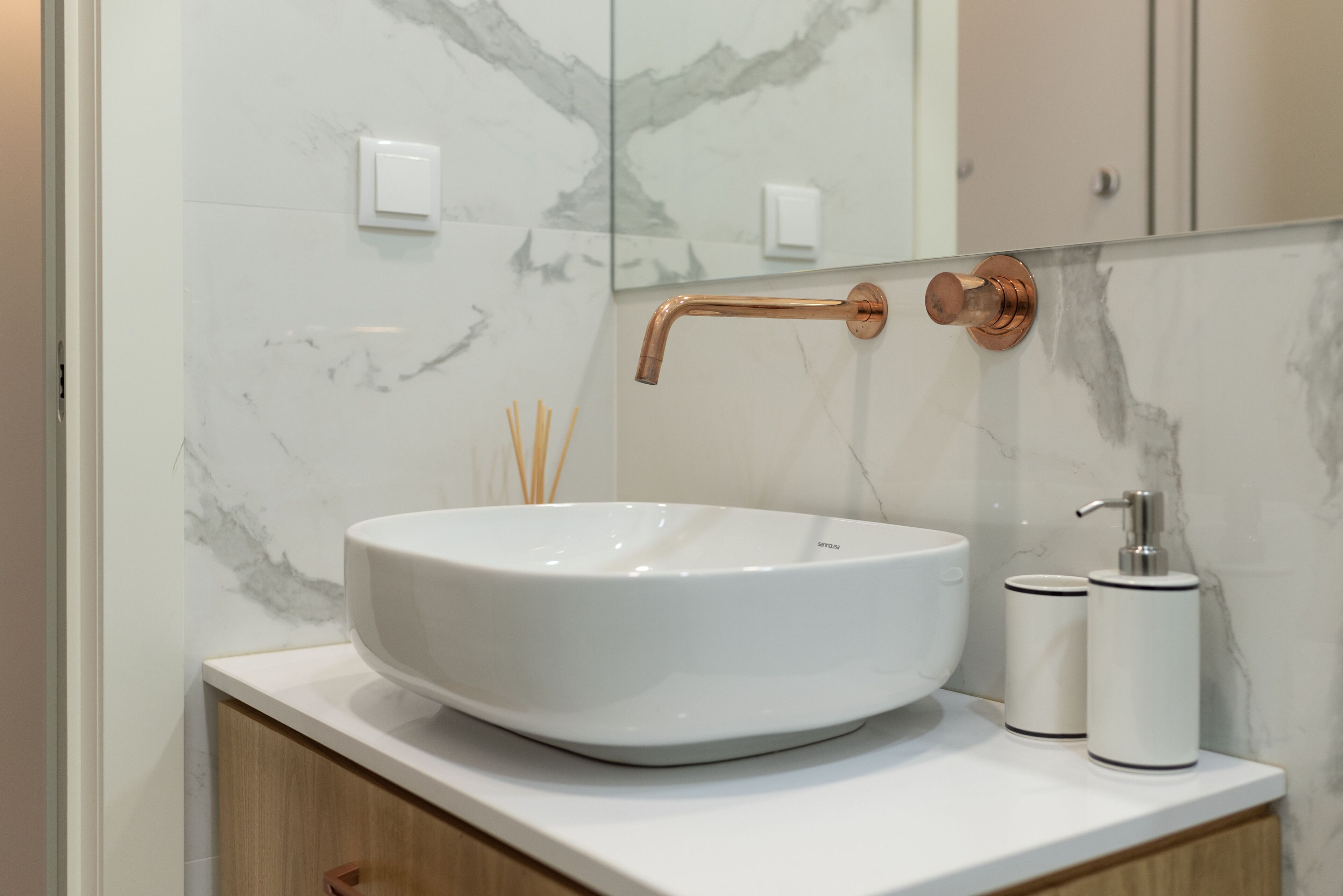 Une salle de bain avec un lavabo en marbre blanc, un robinet en cuivre et deux distributeurs blancs posés sur un comptoir blanc.