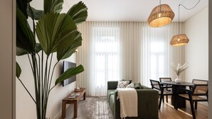 Una sala de estar con un sofá verde, una mesa de comedor con sillas, una planta grande en maceta y dos lámparas colgantes.