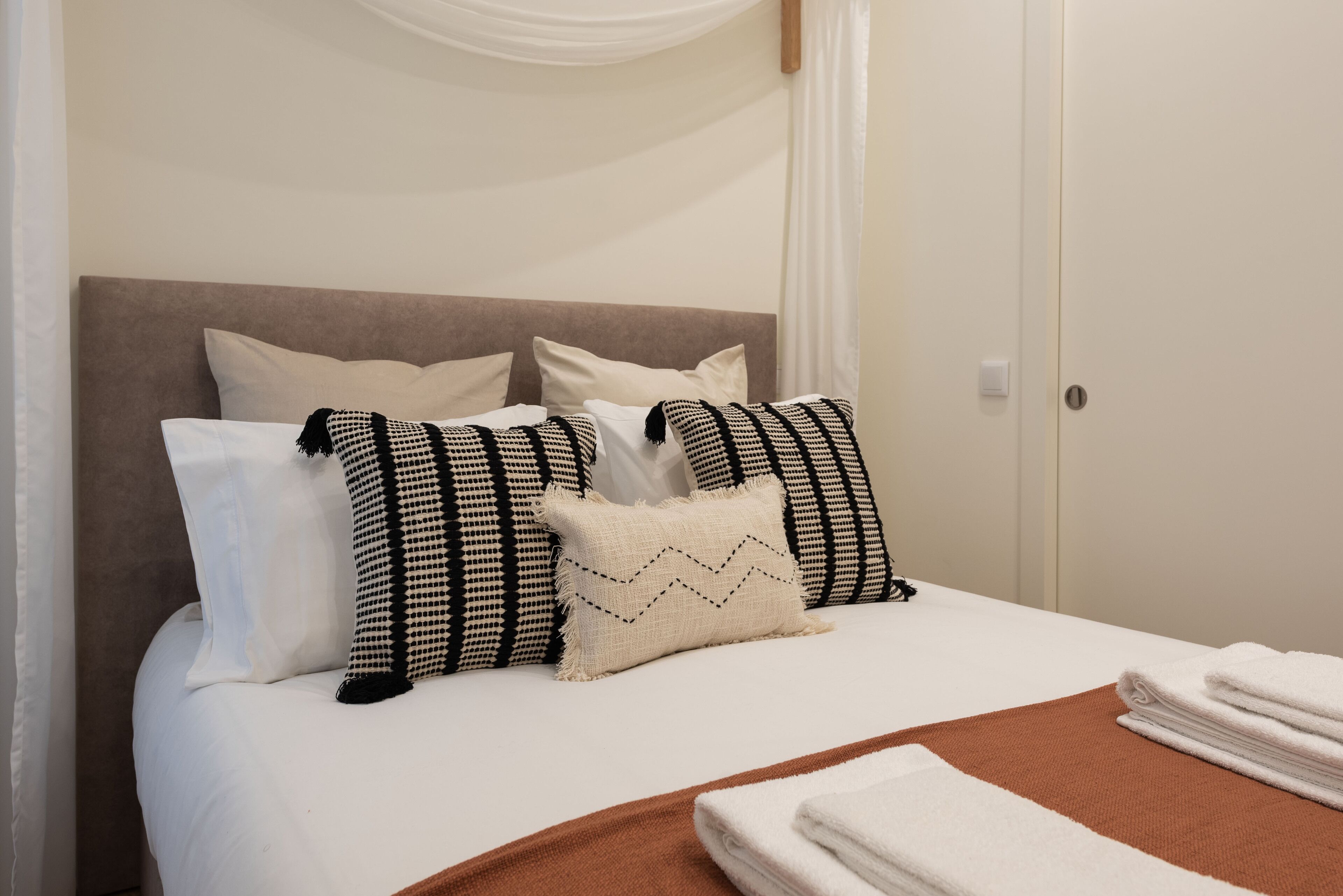 A neatly made bed with white linens and a brown throw blanket, accompanied by patterned pillows and a folded white towel on the side.