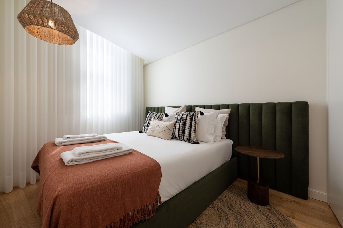 A neatly made bed with a green headboard, white linens, and a brown throw blanket. There is a small round wooden table beside the bed.